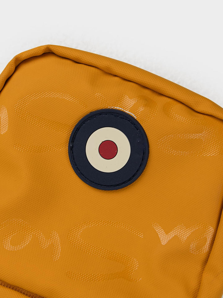 Close-up of Ben Sherman Compact Crossbody Bag in Dijon showing Mod target emblem badge and wax plastisol branding detail