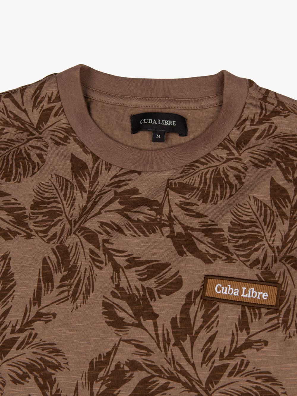 Close-up of Cuba Libre Tee in Beige showing all-over botanical leaf print detail and woven logo patch at chest