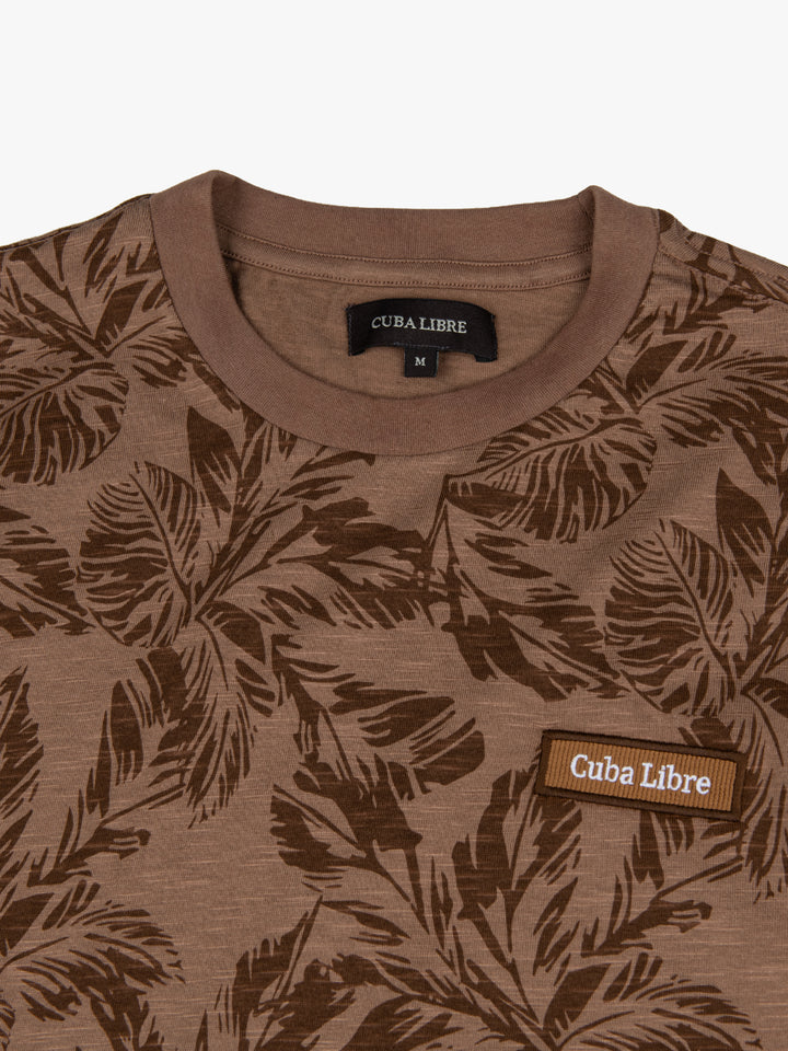 Close-up of Cuba Libre Tee in Beige showing all-over botanical leaf print detail and woven logo patch at chest