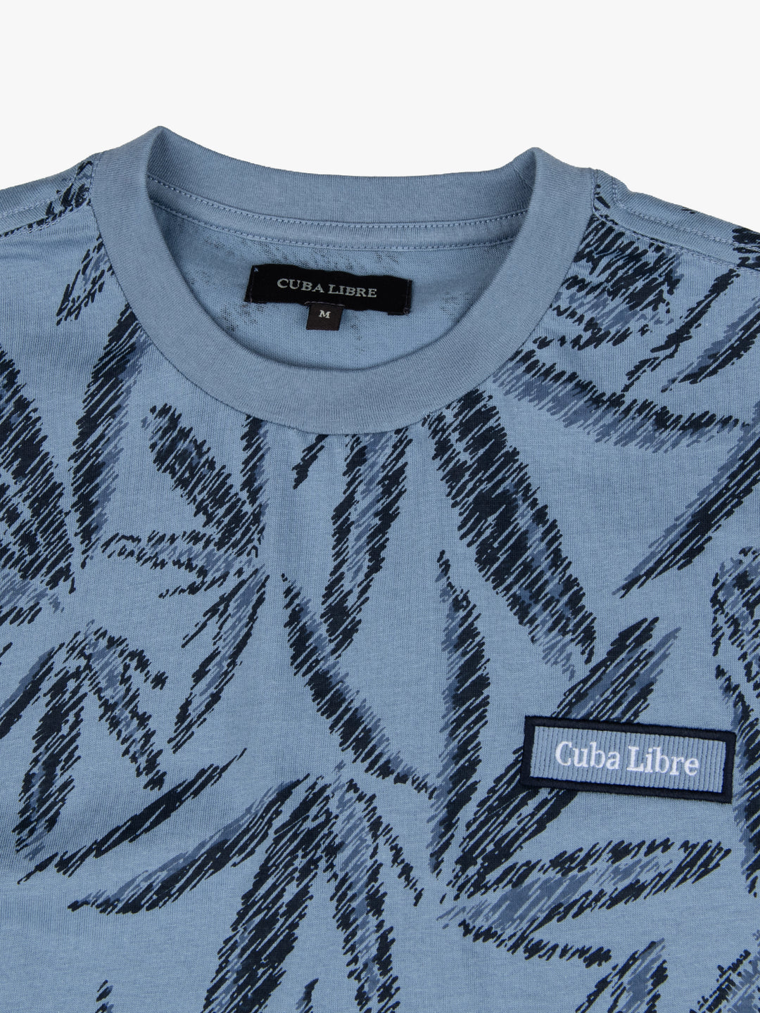 Close-up of Cuba Libre Tee in Dove Blue showing all-over leaf print detail and woven logo patch at chest