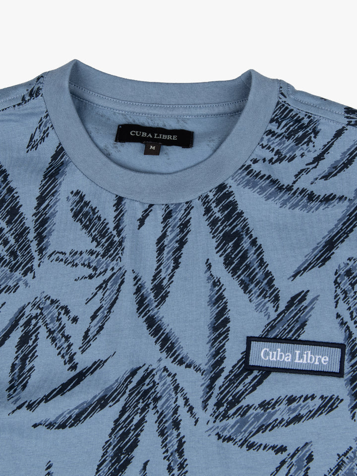 Close-up of Cuba Libre Tee in Dove Blue showing all-over leaf print detail and woven logo patch at chest