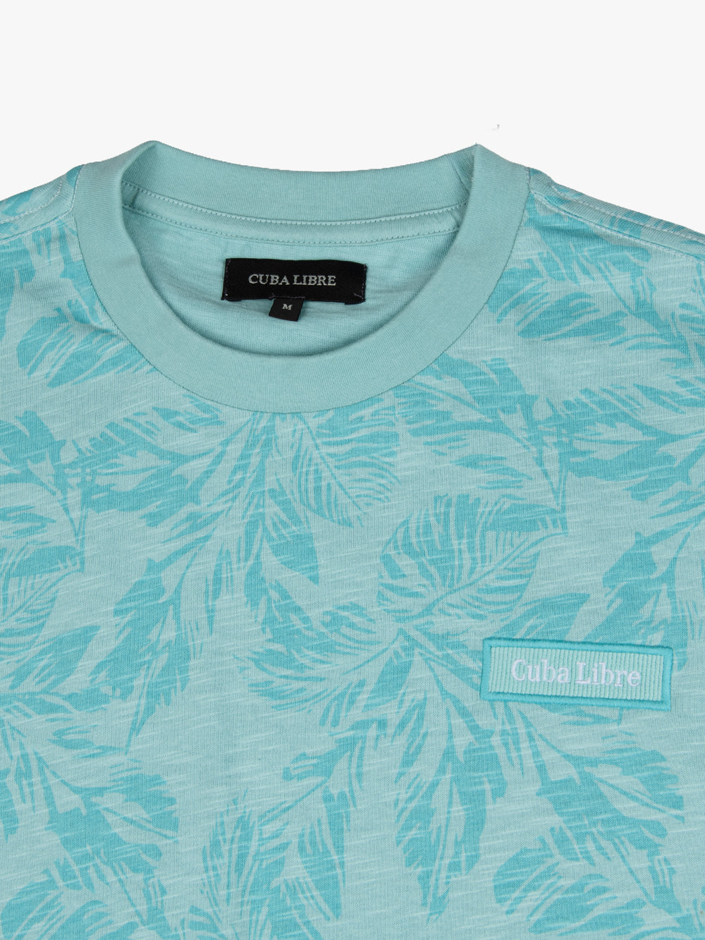 Close-up of Cuba Libre Tee in Turquoise showing all-over leaf print and woven logo patch at chest