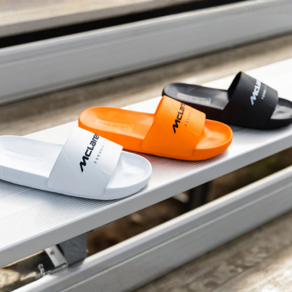 Three McLaren-branded slide sandals in white, orange, and black, arranged on metal stadium-style steps.